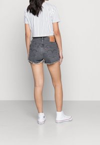 Kobieta ubrana w czarne szorty dżinsowe, białą bluzkę w paski o kroju cropped i białe sneakersy typu high-top, stojąca na jednolitym tle.