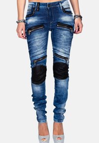 Jeans in denim blu aderenti con molte cerniere, toppe nere sulle ginocchia e dettagli usurati. Presentano accessori in argento e un design affusolato.