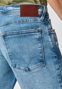 Vaqueros de mezclilla azul claro con un parche de cuero marrón en la cintura, que presentan costuras contrastantes y un bolsillo trasero con un diseño sutil.