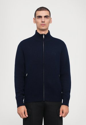 Navy zip-up sweater with a high collar, made of knit fabric. Features two side pockets with zippers and subtle logo detail.