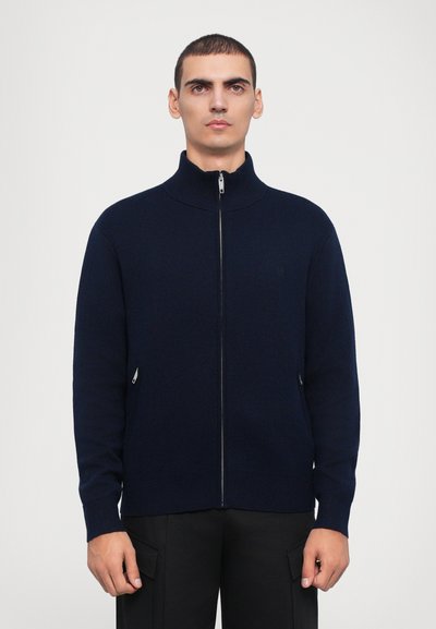 Granatowy sweter zapinany na zamek z wysokim kołnierzem, wykonany z dzianiny. Posiada dwie boczne kieszenie z zamkami błyskawicznymi oraz subtelny detal z logo.