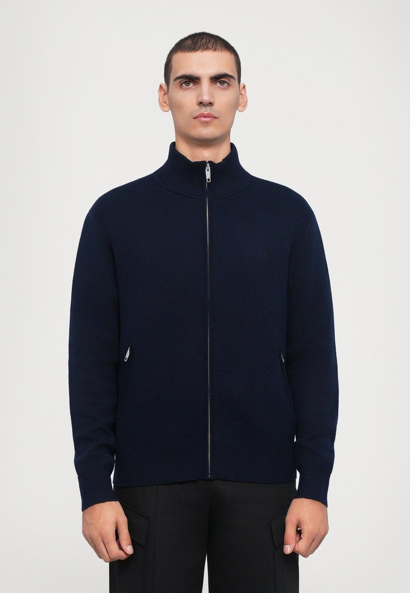 Suéter de punto navy con cremallera y cuello alto. Cuenta con dos bolsillos laterales con cremalleras y un discreto detalle del logotipo.