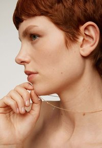 Collar de cadena dorada con pequeños charms geométricos plateados, sostenido por una persona cerca de su mentón, mostrando su delicado diseño.