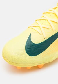 Bota de fútbol amarilla con textura de malla, que presenta un chispeante swoosh verde de Nike y tachuelas naranjas en la suela, diseñadas para proporcionar tracción.