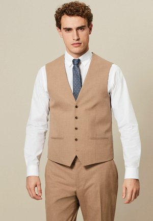 Gilet marron avec une finition texturée, doté de cinq boutons, de poches latérales et d'une coupe ajustée, associé à une chemise blanche et une cravate à motifs.