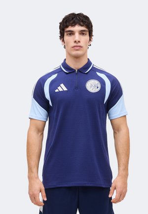 Muškarac u polo majici tamnoplave i svijetloplave boje kratkih rukava sa zatvaračem i sportskim logotipima, stoji na jednostavnoj svijetloj pozadini.