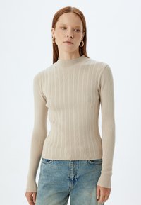 Beżowy sweter prążkowany z wysokim kołnierzem, długimi rękawami i dopasowanym krojem. Noszony z jasnoniebieskimi dżinsami, z drobnym odbarwieniem.