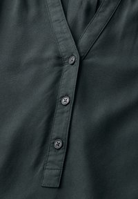 Chemisier vert foncé à col en V, comportant cinq boutons sur la patte, fabriqué en tissu lisse avec un léger éclat.