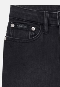 Jeans de denim negro con un ajuste ceñido, acentos de hardware plateado y una etiqueta de la marca Calvin Klein en el bolsillo delantero. Textura lisa.