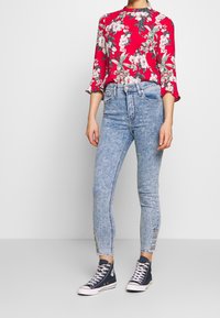 Blusa roja con estampado floral y mangas con volantes, combinada con vaqueros ajustados de tiro alto y lavado claro, con detalles de botones en el tobillo. Zapatillas negras.