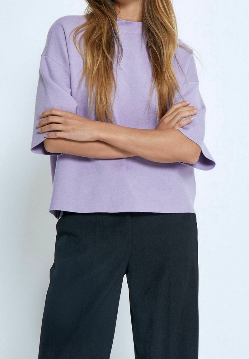 Personne aux longs cheveux portant un haut violet clair à manches courtes et un pantalon noir, debout, bras croisés, devant un fond uni.