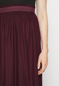 Le bras droit d'une personne portant une jupe violet foncé avec une ceinture texturée et un haut noir, sur fond blanc.