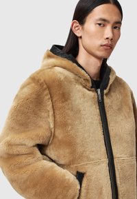 Hellbeige Kunstfelljacke mit schwarz gefütterter Kapuze und Frontreißverschluss. Hat eine boxy Form und Seitentaschen; weiche Textur mit sichtbaren, plüschigen Fasern.