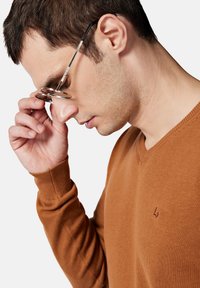 Brązowy sweter w serek wykonany z miękkiego materiału, z ribbowanymi mankietami i małym logo wyszytym na piersi, w zestawieniu z okrągłymi okularami.