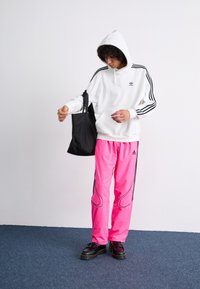 Sweatshirt branco com detalhe de três riscas pretas, combinado com calças de atletismo rosa brilhante com pormenores pretos. Bolsa de ombro preta e sapatos.