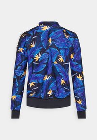 Veste avec imprimé feuilles tropicales bleues et fleurs jaunes, poignets et taille noirs, et texte de la marque en blanc sur fond foncé.