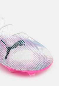 Scarpino da calcio bianco con una parte superiore testurizzata, caratterizzato da un motivo a gradiente di punti blu e rosa, e una suola traslucida rosa. Logo nero visibile.