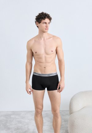 Črne boksarice s sivo elastično pasom. Material je videti gladek in raztegljiv. Model stoji na svetli teksturirani površini.