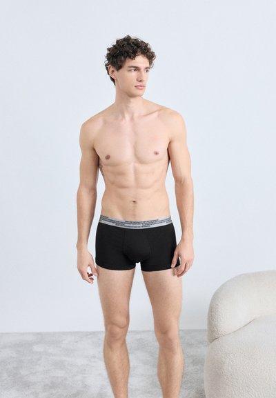 Boxer neri con fascia elastica grigia in vita. Il materiale sembra liscio ed elastico. Il modello è in piedi su una superficie chiara e testurizzata.