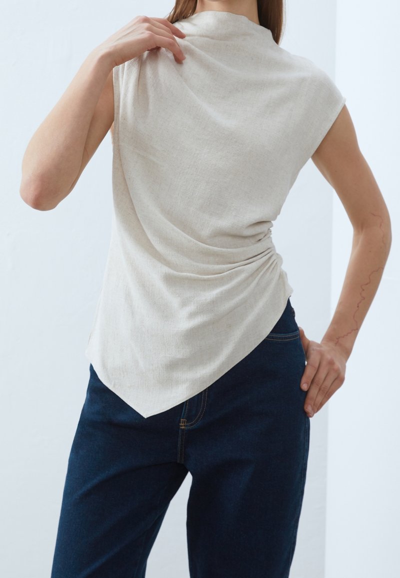 Haut beige sans manches avec texture drapée, encolure large et ourlet asymétrique, associé à un jean bleu foncé pour le contraste.