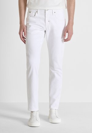 Hombre con jeans blancos ajustados y zapatillas blancas de pie contra un fondo claro sencillo.