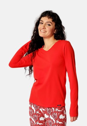 Frau mit langen, lockigen, dunklen Haaren, lächelnd, trägt ein rotes Langarmshirt und gemusterte rot-weiße Hosen vor einem einfarbigen Hintergrund.
