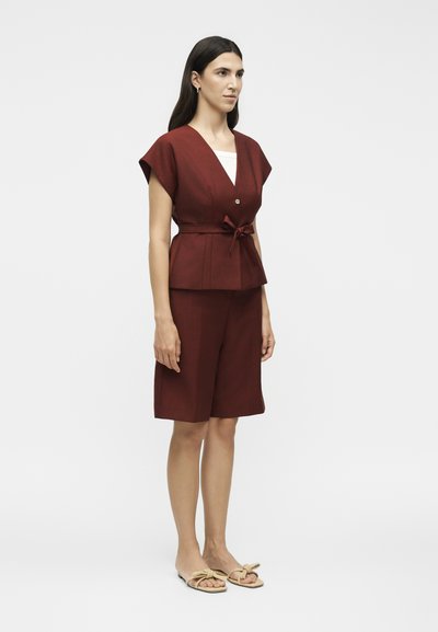 Γυναίκα που στέκεται φορώντας κορυφή σε σκουριά απόχρωση με κοντά μανίκια και ζώνη, καθώς και σορτς μέχρι το γόνατο στο ίδιο χρώμα και μπεζ σανδάλια τύπου slip-on με φιόγκο.