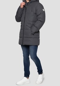 Dunkelgraue Steppjacke mit Kapuze, ausgestattet mit einem Frontreißverschluss, zwei seitlichen Taschen und einer Brusttasche. Getragen über blauen Jeans und weißen Sneakers.
