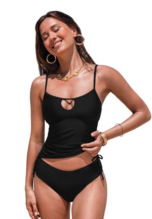 Donna con tankini nero con dettaglio a occhiello, orecchini a cerchio dorati, collana e braccialetti, sorridente a occhi chiusi su sfondo bianco.
