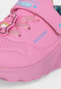 Chaussure pour enfant Skechers rose avec bride Velcro, lacets rose clair et breloque ours dessin animé près des orteils.