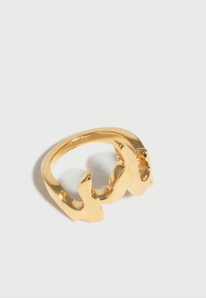 Δαχτυλίδι από μεταλλικό υλικό με χρυσή απόχρωση και λείο, κυματιστό σχέδιο που ακουμπά σε λευκή επιφάνεια.