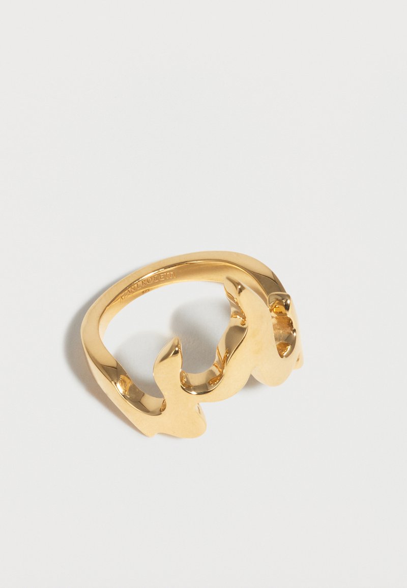 Δαχτυλίδι από μεταλλικό υλικό με χρυσή απόχρωση και λείο, κυματιστό σχέδιο που ακουμπά σε λευκή επιφάνεια.