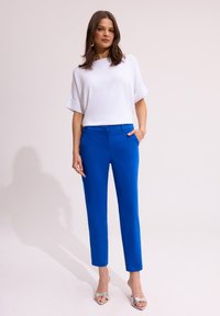 Biała bluzka z krótkim rękawem o luźnym kroju zestawiona z eleganckimi, jasnoniebieskimi spodniami. Modelka nosi srebrne sandały na obcasie odkryte z przodu oraz dodatki.
