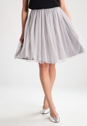 Femme portant une jupe plissée en tulle gris clair, un haut noir et des ballerines argentées, debout, jambes croisées sur fond blanc.