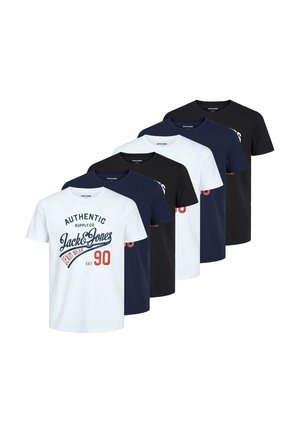 Man wearing white graphic t-shirt, dark blue jeans, and white sneakers, standing with hands loosely clasped in front against plain background.