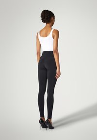 Leggings neri vita alta in tessuto elasticizzato, silhouette aderente, abbinati a scarpe nere con tacco alto, modello visto da dietro.