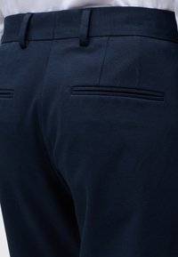 Κοντινή λήψη της πίσω όψης σκούρων ναυτικών επίσημων παντελονιών με δύο οριζόντιες τσέπες και θηλιές για ζώνη, φορεμένα με μια λευκή πουκάμισο που είναι μέσα στην παντελόνα.