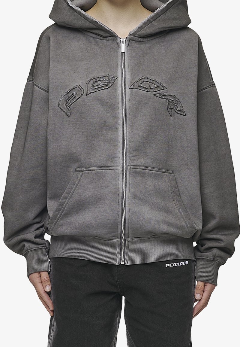 Personne portant un sweat à capuche gris zippé avec une couture abstraite sur la poitrine et les poches avant, associé à un pantalon noir étiqueté "PEGADOR."