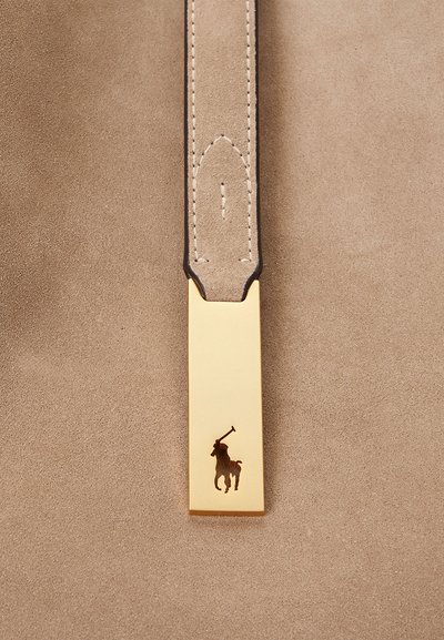 Auksinės detalės su įrėžtu polo žaidėjo siluetu, pritvirtintos prie šviesiai rudos zomšos dirželio, pasižyminčio tvarkingu siuvimu ir lygiu paviršiumi.
