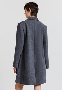 Cappotto grigio di lana con vestibilità sartoriale, maniche lunghe e colletto a rever. Caratterizzato da un design pulito e un tessuto testurizzato, arriva fino a metà coscia.