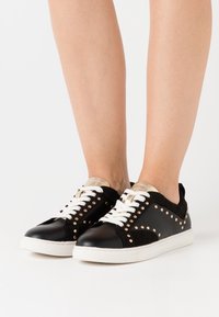 Svarta läder- och mockaskor med guldaccenter, nitar, vita snören och en platt gummisula. Inga distinkta mönster.