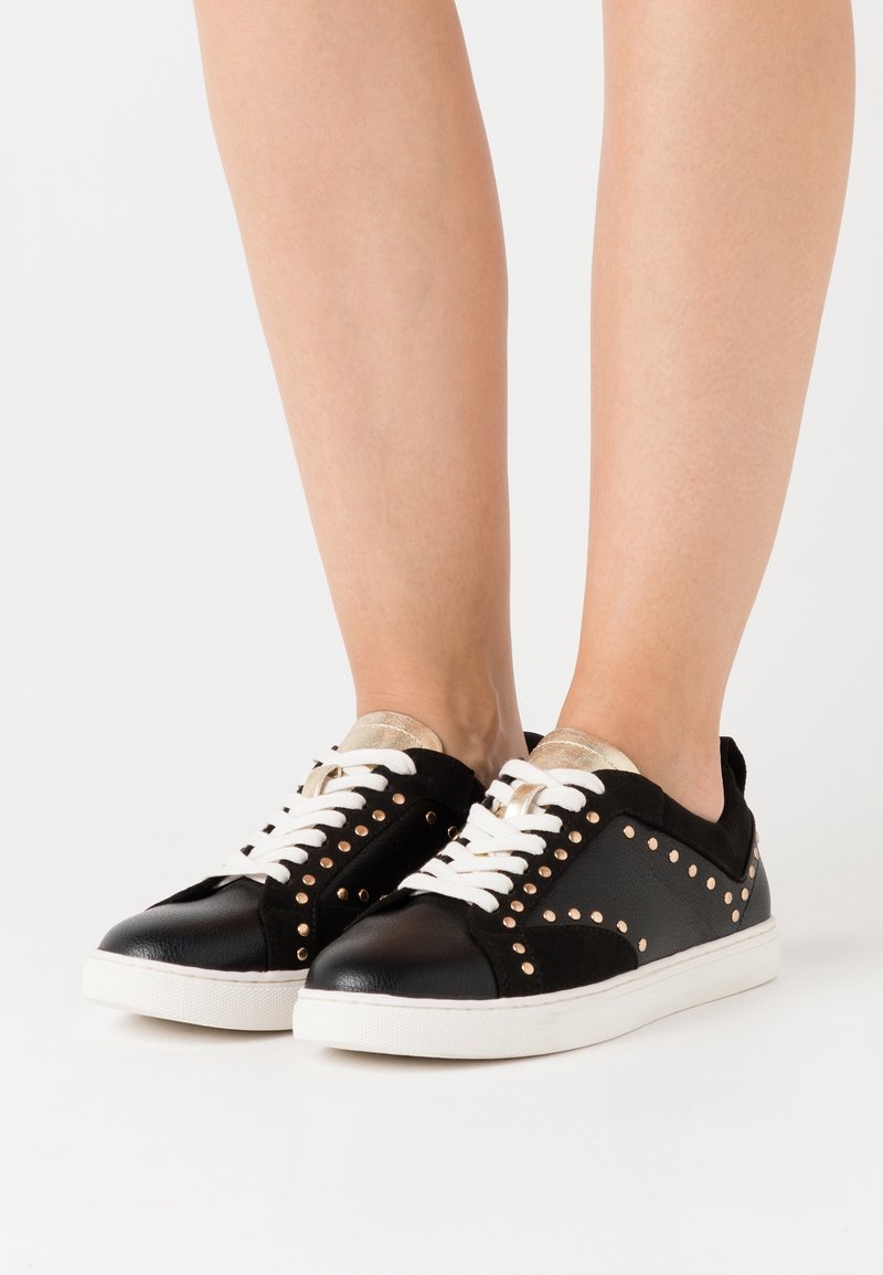 Svarta läder- och mockaskor med guldaccenter, nitar, vita snören och en platt gummisula. Inga distinkta mönster.