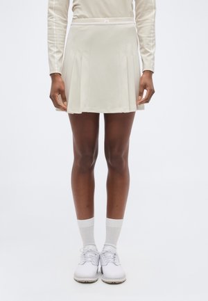 Kvinde iført en lysebeige plisseret nederdel, langærmet top, hvide ankelsokker og hvide sportssko, stående foran en ensfarvet baggrund.