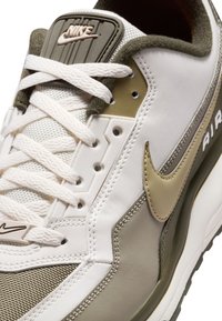 Zapatilla Nike con parte superior sintética blanca, acentos en verde oliva, malla textil, logo prominente y área de cordones texturizada. Suela de goma visible.