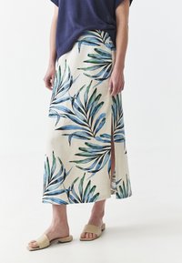 Jupe maxi florale en tissu naturel avec des motifs de feuilles vertes et bleues, dotée d'une fente latérale et associée à des sandales à glissière beiges.