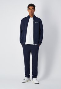 Completo da ginnastica navy realizzato in tessuto morbido, composto da giacca con zip e pantaloni abbinati. Maglietta bianca sotto e accenti del logo visibili.