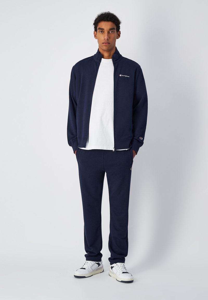 Completo da ginnastica navy realizzato in tessuto morbido, composto da giacca con zip e pantaloni abbinati. Maglietta bianca sotto e accenti del logo visibili.