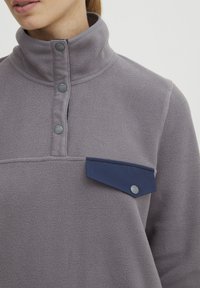 Pullover in pile grigio chiaro con colletto alto, dotato di bottoni grigi e una tasca a pattina blu marina sul petto. Texture morbida.