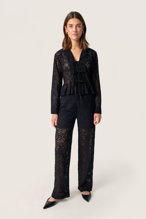 Black lace top and pants set featuring a peplum waist, V-neck with tie detail, and long sleeves. Finished with pointed black shoes.