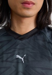 Tuvumā redzams cilvēka kakls un apkakle, kas valkā tumšu rakstainu sporta krekli, uz kura redzams izteikts baltais Puma logotips.